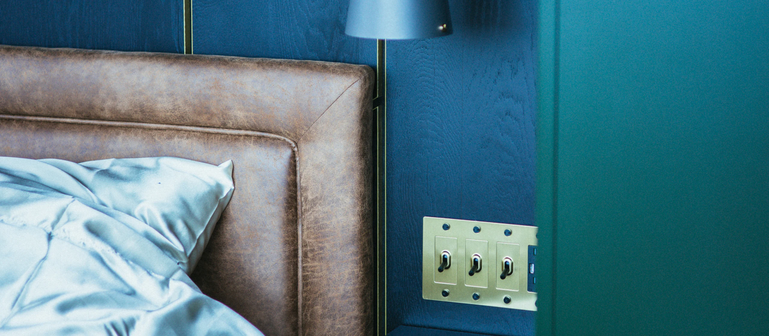 Bedroom detail — tan leather headboard, silk bedding, and brass toggle switches against deep blue-green oak walls