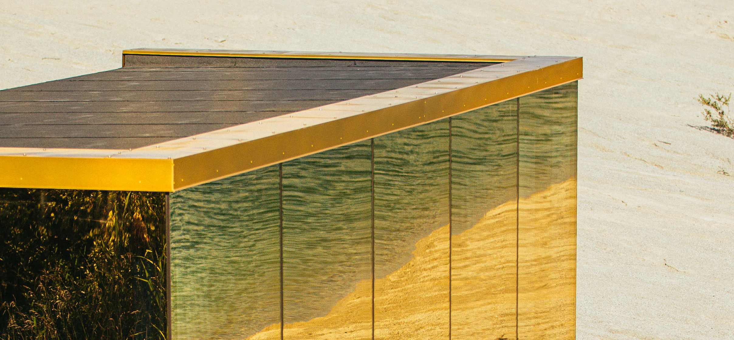 Brass roofline trim meeting mirrored glass panels that reflect the surrounding sand dunes